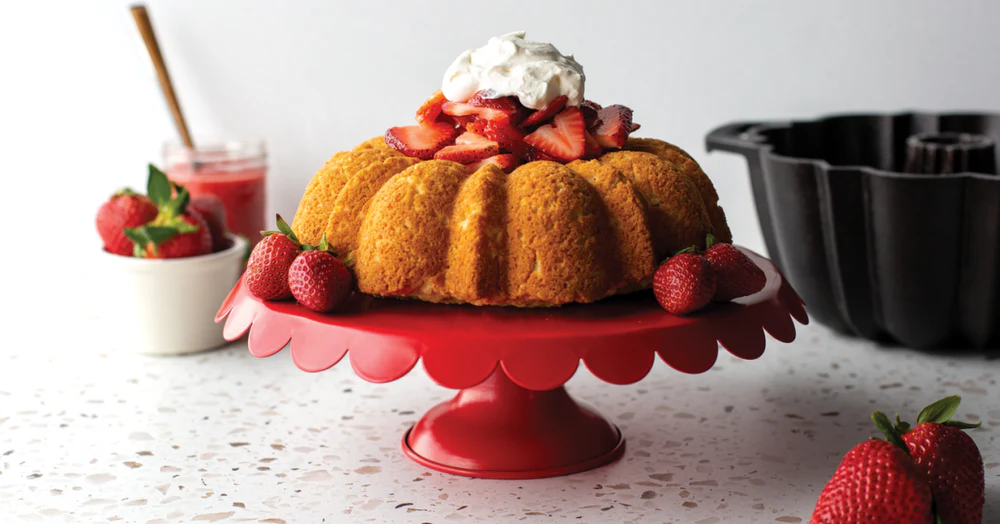 Prhki kolač sa svježim jagodama i šlagom serviran na crvenom stalku pečen u rebrastom kalupu.