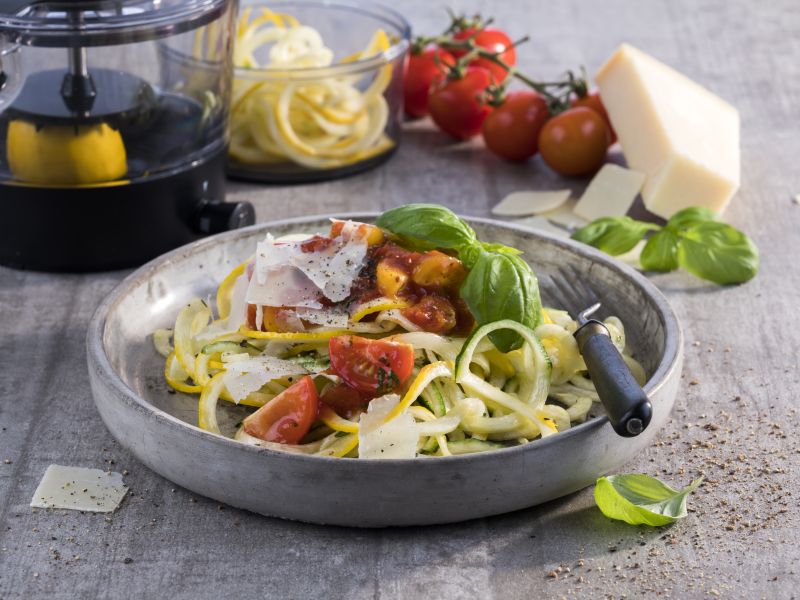 Zdravi domaći špageti od tikvica posluženi na tanjuru uz svježe cherry rajčice i GEFU spiralni rezač u pozadini.